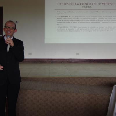 hombre de pie  habla con micrófono en mano y proyecta presentación 