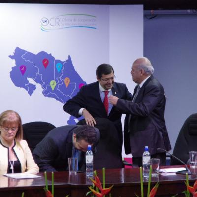 Dos ejecutivos de pie y una mujer de blanco sentada en mesa de reuniones
