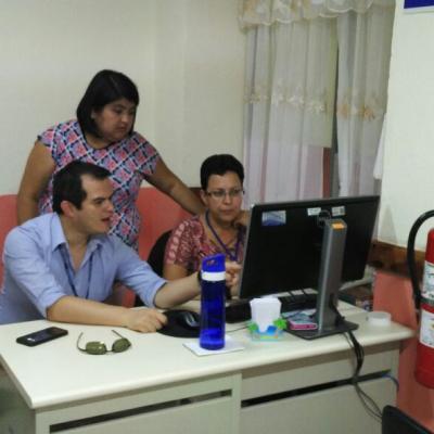 Dos personas sentadas en el escritorio y una de pie señalando 